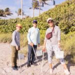 Three people at the beach dressed and holding trash collecting tools