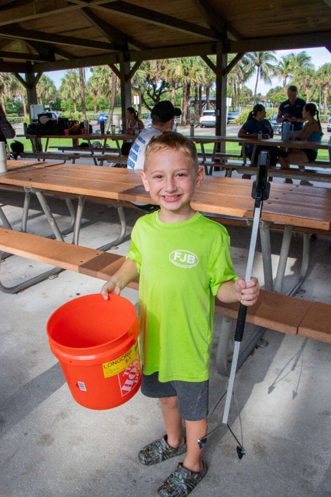 Young beach cleanup volunteer holding up trash collection tools under the Anita Lankler Pavilion in Juno Beach.
