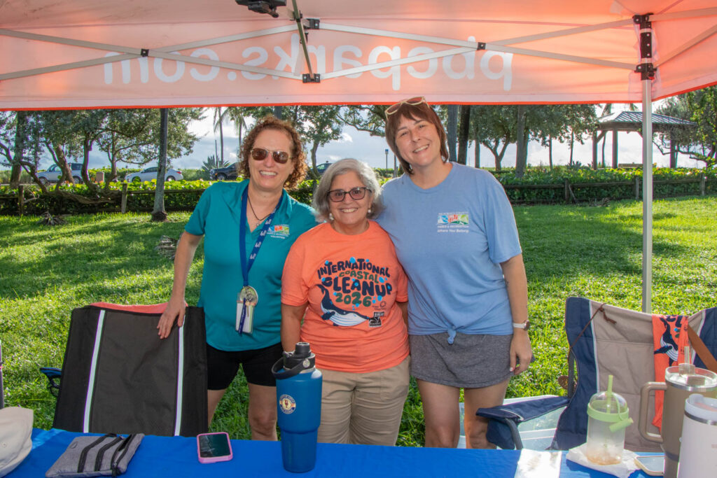 Three volunteers smiling under a tent by Jupiter Beach.