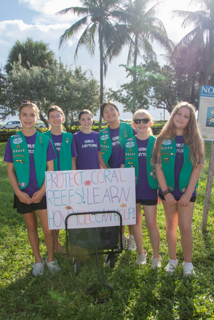 Group of Girl Scouts smiling and standing behind a homemade sign in the grass.