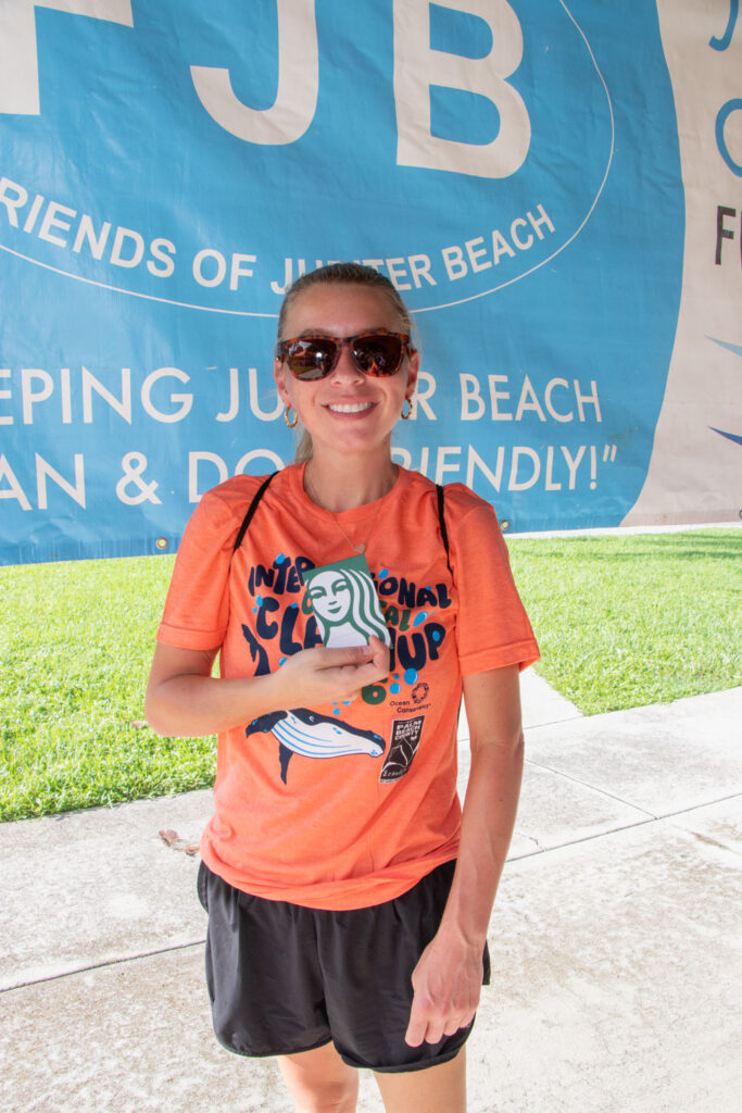 Young female volunteer in orange International Coastal Cleanup shirt holding up a Starbucks Coffee gift card and smiling.