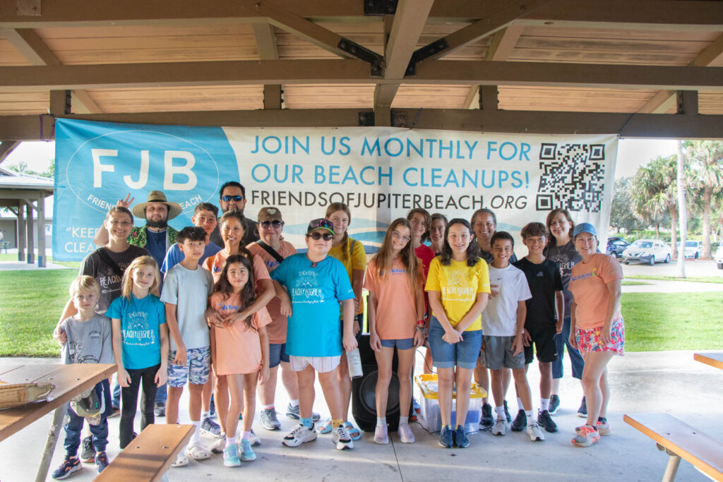 Group of beach cleanup volunteers standing under a pavilion in front of the Friends of Jupiter Beach banner.