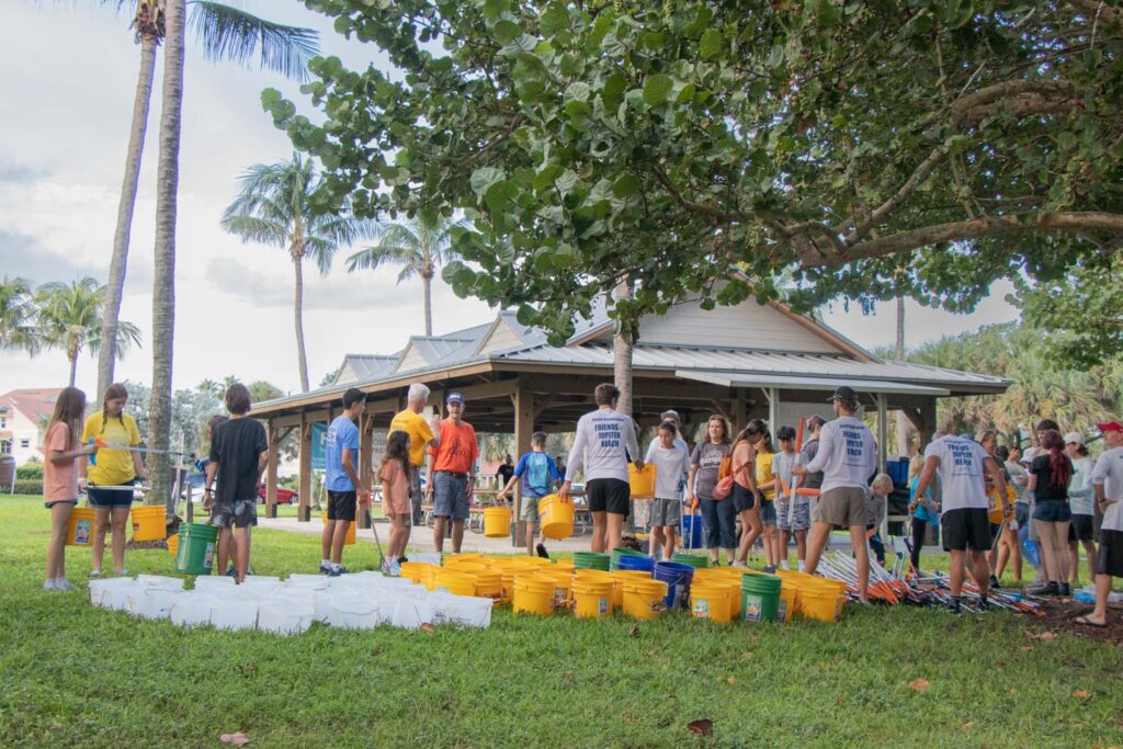 Beach cleanup volunteers holding buckets and cleanup supplies on the grass.