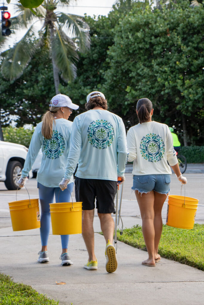 Beach cleanup volunteers holding bucket and cleanup supplies and walking on the sidewalk.