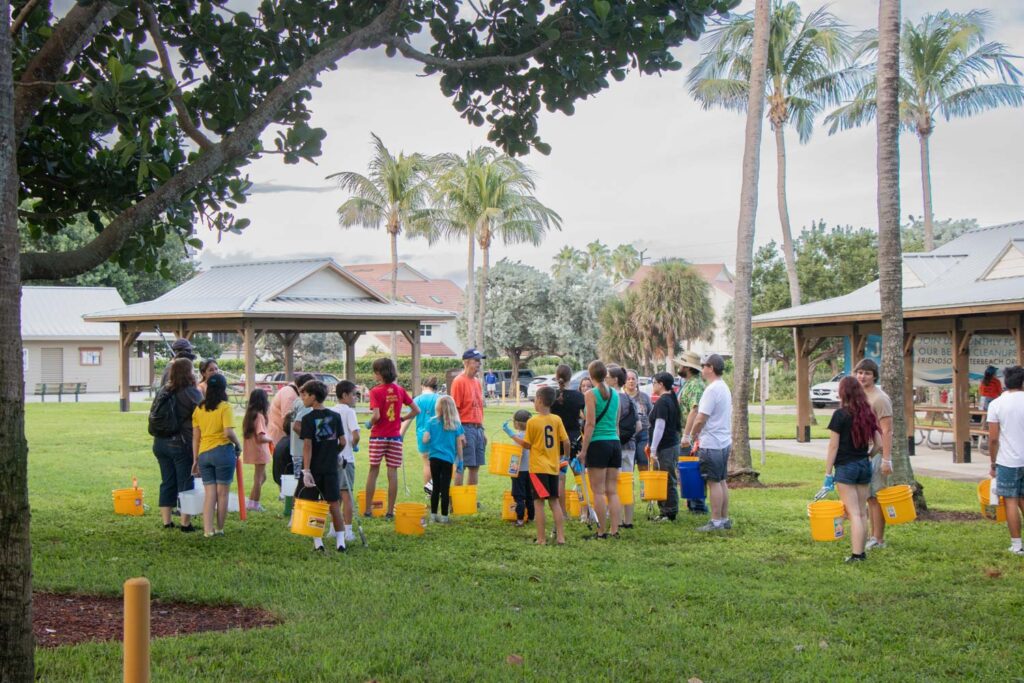 Beach cleanup volunteers holding bucket and cleanup supplies on the grass.