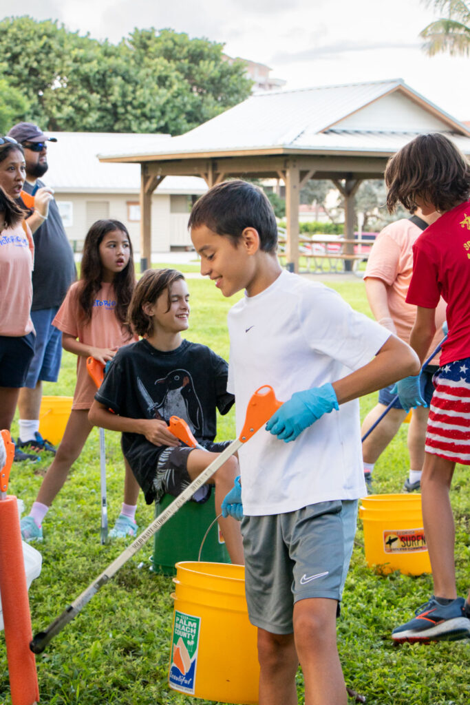 Beach cleanup volunteers holding bucket and cleanup supplies on the grass.