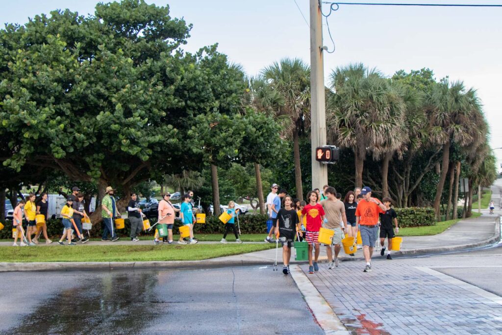 Beach cleanup volunteers holding bucket and cleanup supplies and walking across the street.