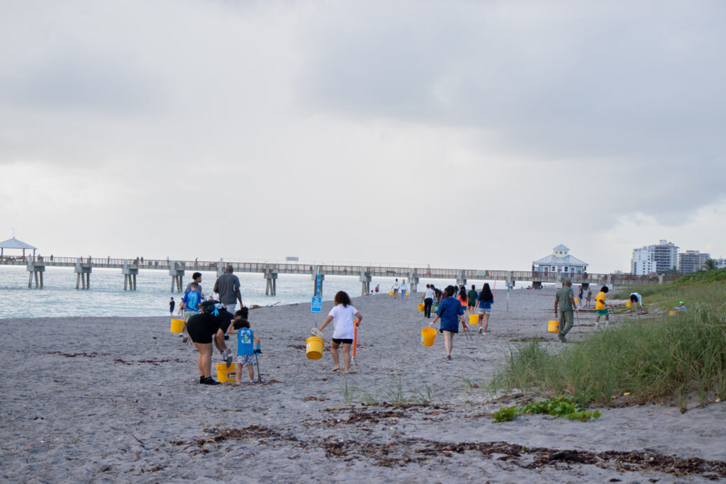 Beach cleanup volunteers holding bucket and cleanup supplies and walking across the beach.