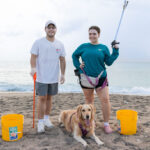 A man, woman, and their dog posing on the beach with cleanup supplies.